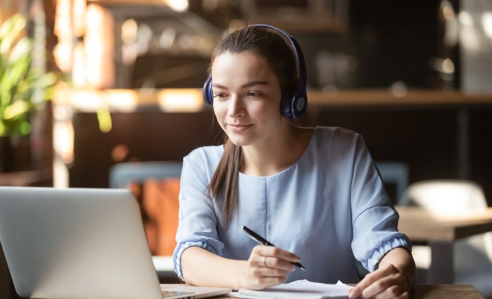 Focused,Woman,Wearing,Headphones,Using,Laptop,In,Cafe,,Writing,Notes,